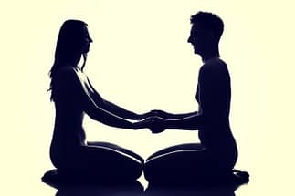 Due persone sedute a gambe incrociate, che si guardano e si tengono per mano in una pratica di sesso tantrico, simbolo di connessione spirituale e intimità profonda.