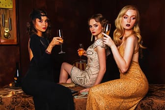 Tre donne raffinate con abiti di lusso e calici di champagne in un’ambientazione esclusiva. Analisi di escort e psicologia - Pommenor.