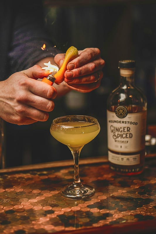 Mani esperte creano un cocktail con tecnica a fiamma, simbolo di mixology raffinata in stile Pommenor.